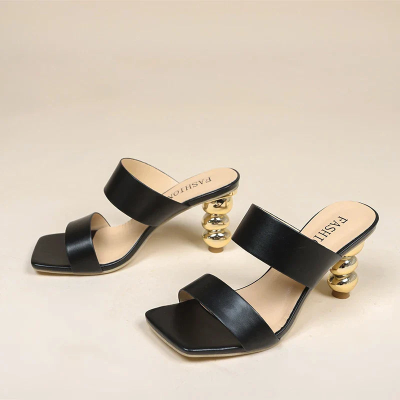 A pair of black open-toe heeled sandals with two wide straps and unique sculpted gold ball-stacked heels, crafted from premium leather and displayed on a neutral background.