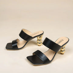 A pair of black open-toe heeled sandals with two wide straps and unique sculpted gold ball-stacked heels, crafted from premium leather and displayed on a neutral background.
