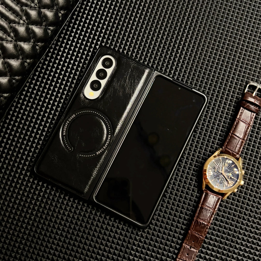 A foldable smartphone in a Samsung Galaxy Z Fold case is placed on a textured black surface next to a brown leather-strap wristwatch with a gold case and blue face. A black quilted item appears in the top left corner.