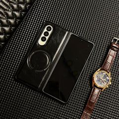 A foldable smartphone in a Samsung Galaxy Z Fold case is placed on a textured black surface next to a brown leather-strap wristwatch with a gold case and blue face. A black quilted item appears in the top left corner.