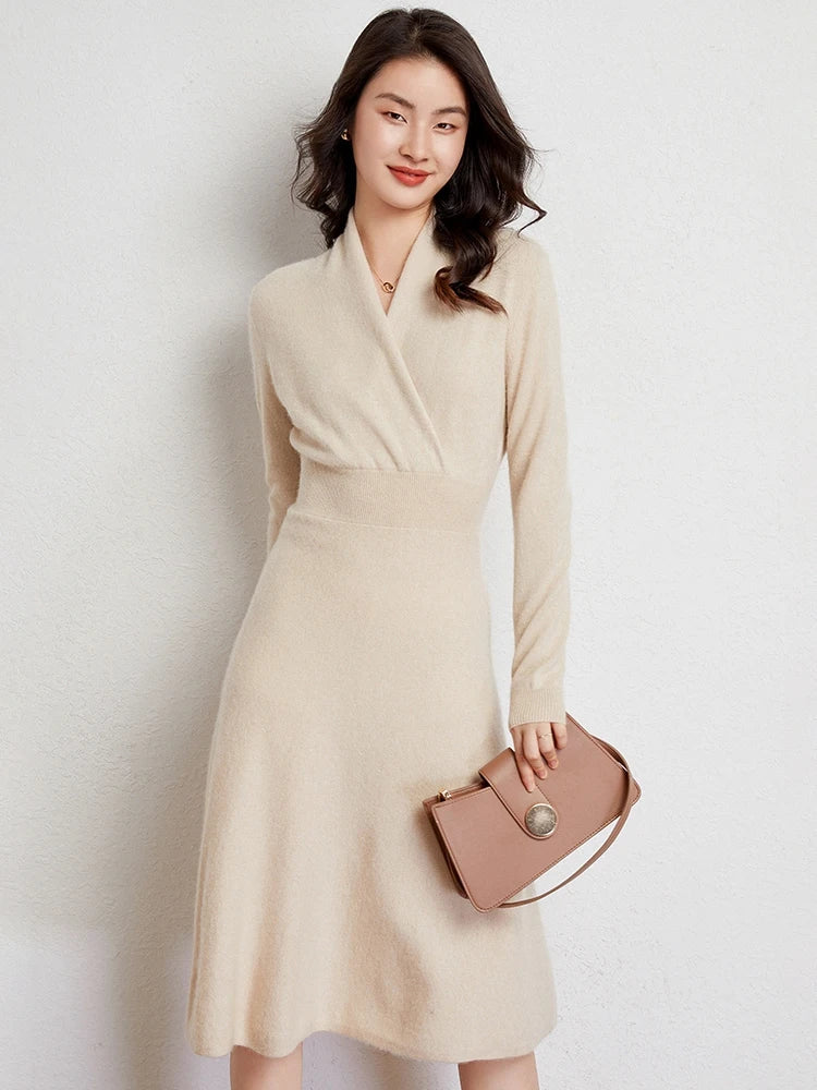 A woman wearing a long-sleeved, cream-colored Matilda Wrap Dress with a V-neck wrap stands against a light wall, smiling, with wavy hair and holding a tan handbag in her left hand.