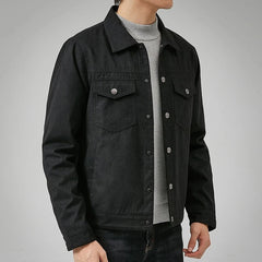 A person in a black button-up jacket with a classic point collar over a light gray sweater and dark jeans stands against a plain gray background, their head partially out of frame.