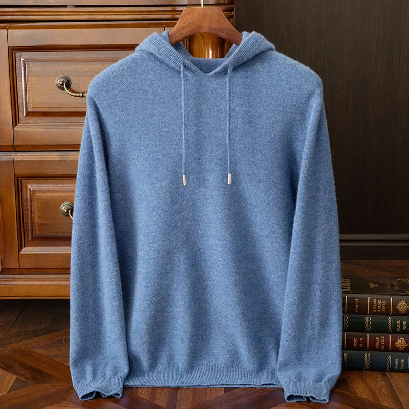 A blue hooded layering sweater with drawstrings hangs on a wooden hanger, displayed in front of a wooden dresser and a stack of books on a polished wood floor.