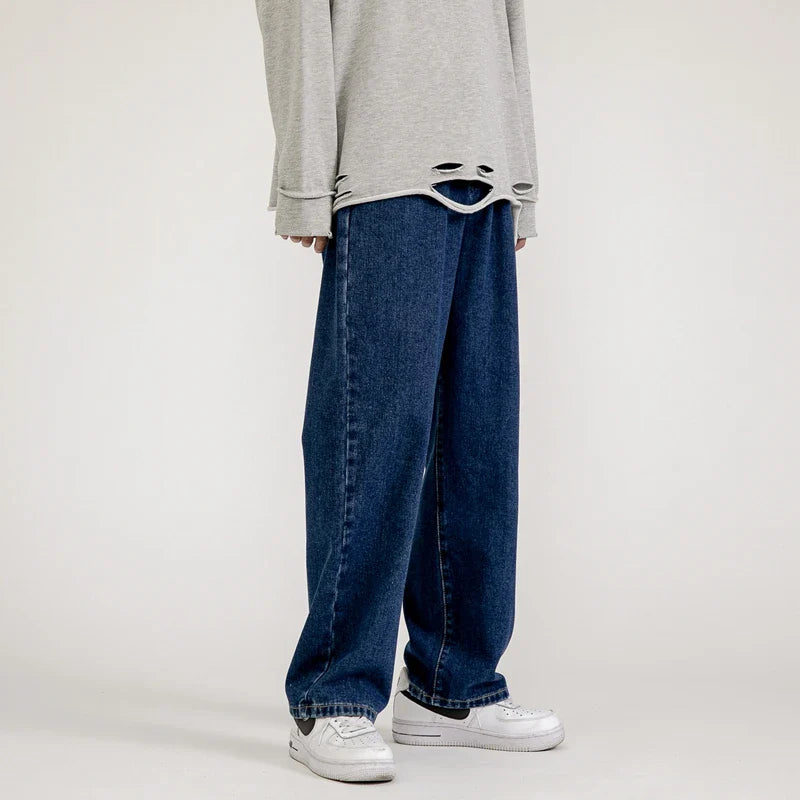 Person wearing a loose, light gray sweater with distressed holes at the hem, relaxed fit jeans in a dark vintage wash, and white sneakers, standing against a plain background.