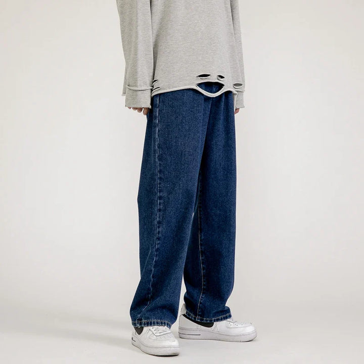 Person wearing a loose, light gray sweater with distressed holes at the hem, relaxed fit jeans in a dark vintage wash, and white sneakers, standing against a plain background.