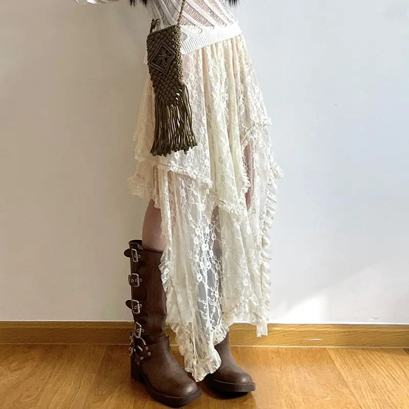 Person wearing a white, high waist asymmetrical lace skirt with a silky lace finish, brown knee-high boots with multiple buckles, and an olive green woven crossbody bag, standing on a wooden floor against a plain white wall.