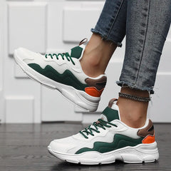 A person wearing advanced shock absorption sneakers in white, green, brown, and orange stands on dark wooden flooring. They are dressed in frayed-hem blue jeans and an anklet, with their feet lifted slightly off the ground.