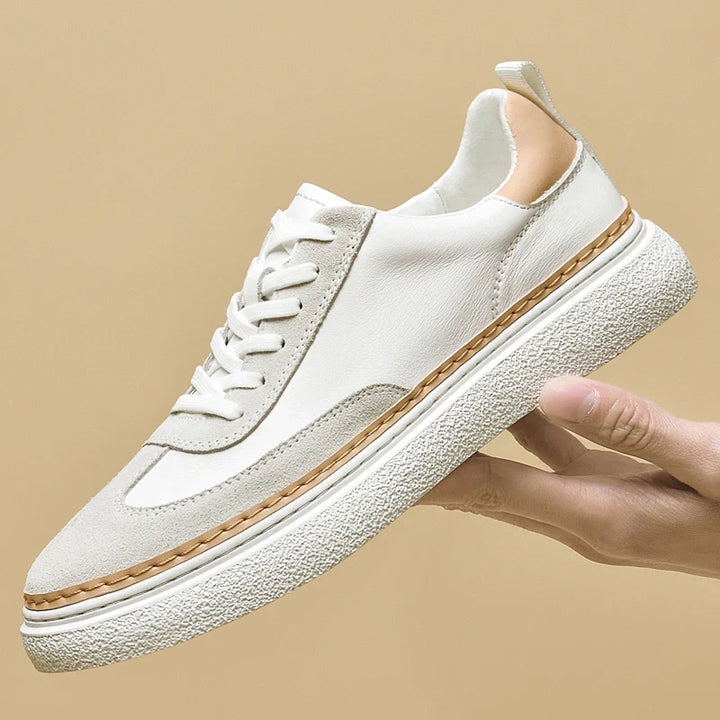 A hand holds up a white sneaker with light beige accents, a textured white sole, and tan stitching details against a plain beige background, highlighting its cushioned insole for extra comfort.