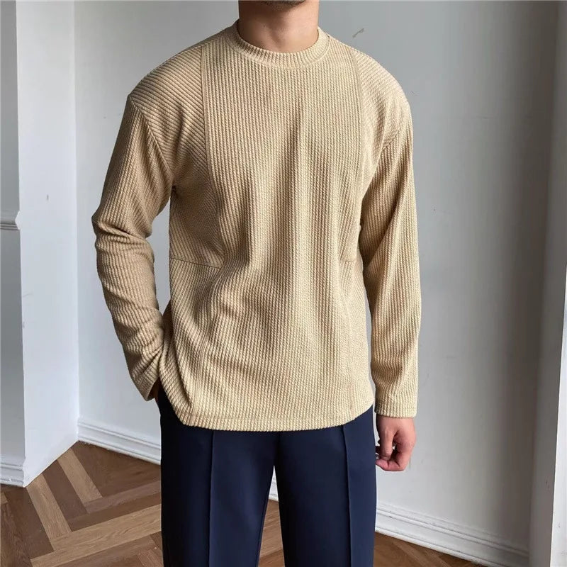 A person wearing a beige, long-sleeve Grayson Crew Classic sweater in premium cotton blend and dark navy pants stands indoors on a wooden floor with one hand in their pocket. The face is not visible.