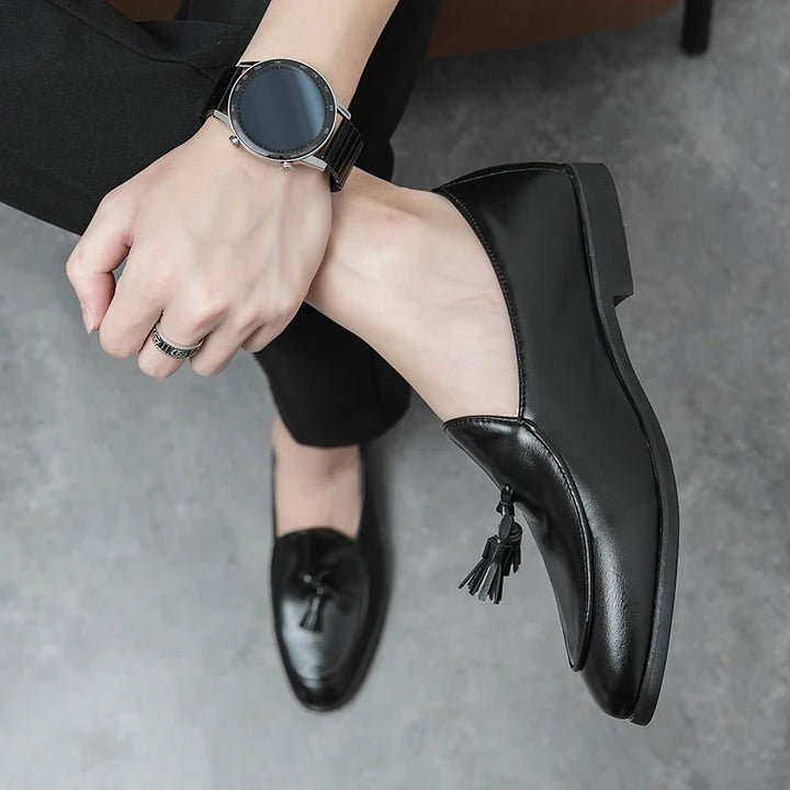 Person wearing black dress pants, black leather tassel loafers with a cushioned insole, a black watch, and a silver ring sits with legs crossed on a gray floor, showcasing the shoes’ vintage look and stylish accessories.