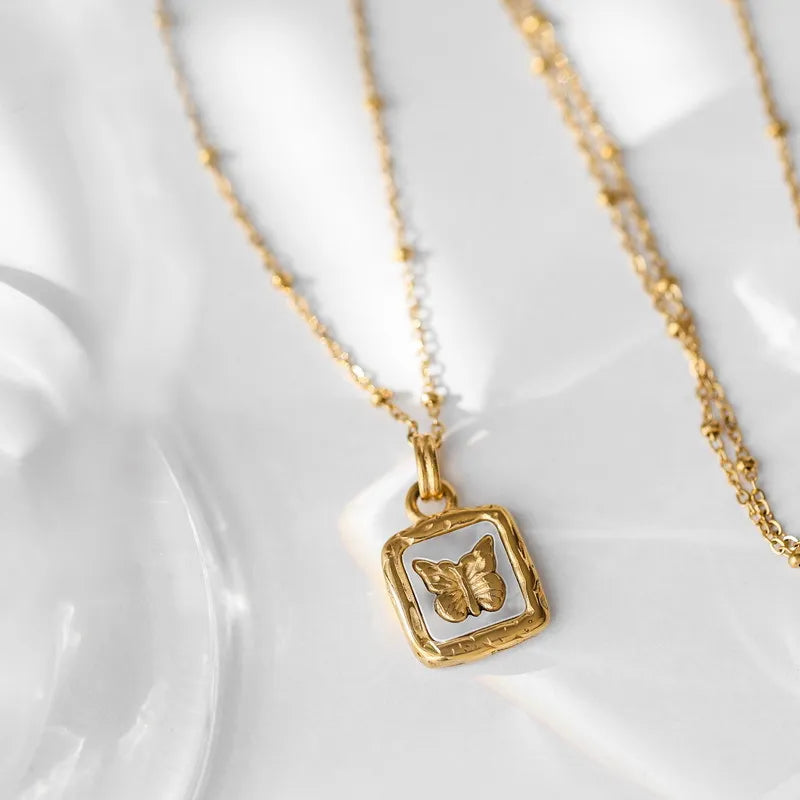 A delicate gold necklace with a square pendant featuring a raised butterfly design, crafted with elegant craftsmanship and displayed on a light, textured surface with soft shadows.