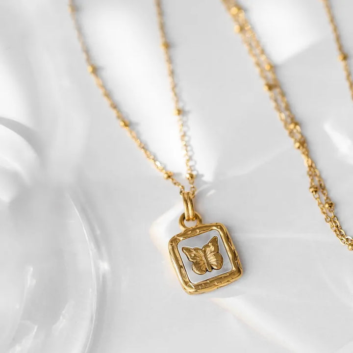 A delicate gold necklace with a square pendant featuring a raised butterfly design, crafted with elegant craftsmanship and displayed on a light, textured surface with soft shadows.