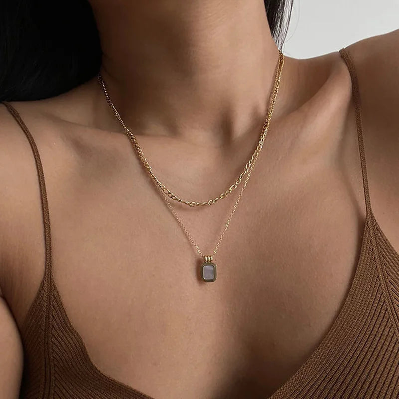 A woman wearing a brown ribbed tank top and two gold necklaces—one is a simple chain, and the other features a rectangular pendant with a light-colored crystal rhinestone.