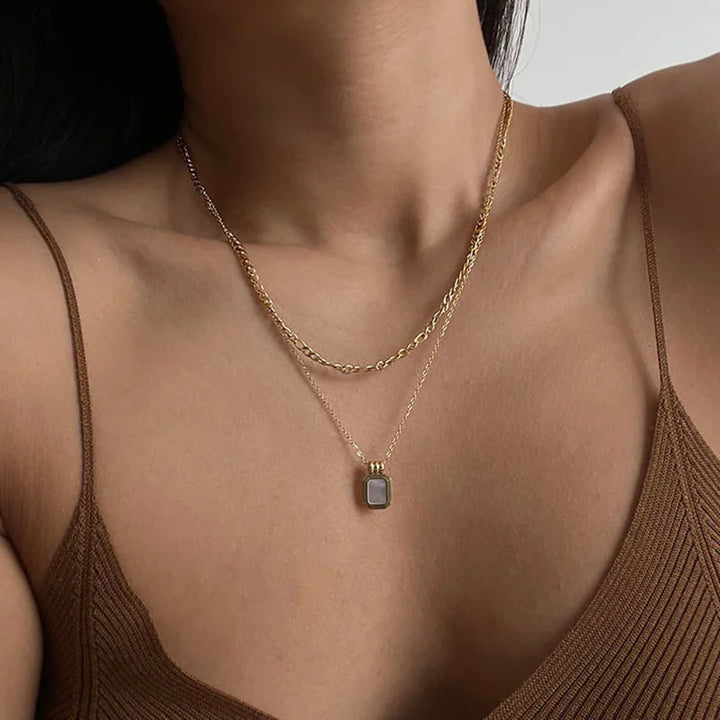 A woman wearing a brown ribbed tank top and two gold necklaces—one is a simple chain, and the other features a rectangular pendant with a light-colored crystal rhinestone.
