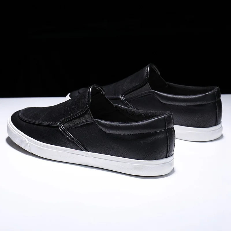 A pair of mens slip-on black casual shoes with a leather insole and white rubber-leather sole are displayed on a white surface against a black background.