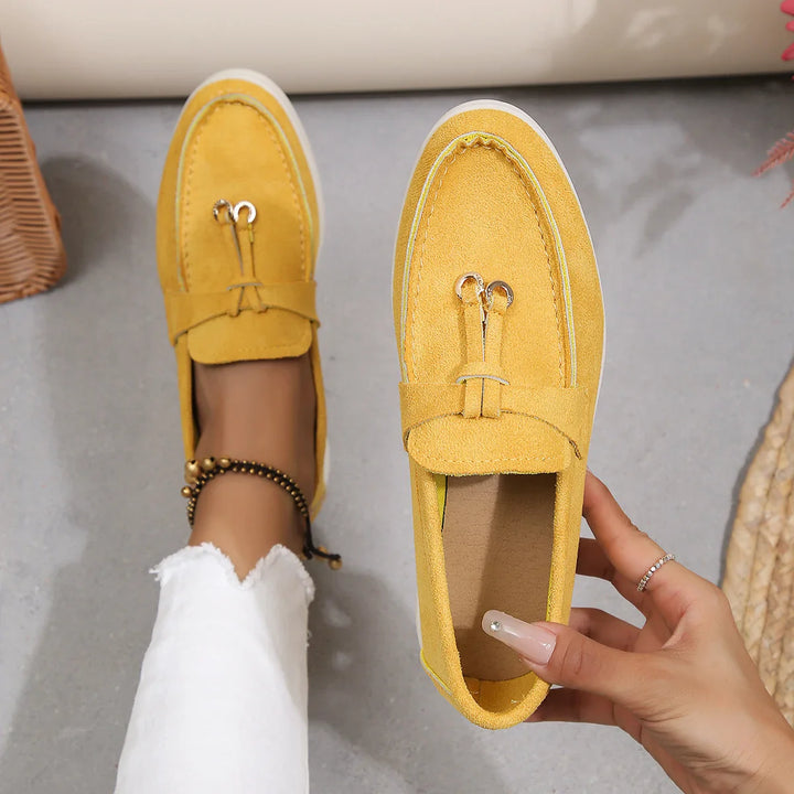 A person wearing white pants and a beaded anklet tries on yellow suede moccasins with a metal detail, holding one shoe in hand while the other is on their foot, against a light gray background.