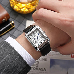 A person in a gray pinstripe suit wears a luxury watch with a square silver case, white dial, black Roman numerals, and premium leather band. A glass of whiskey, pen, and newspaper rest in the background.
