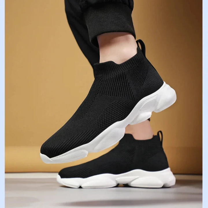 A person wearing black slip-on athletic sneakers with white chunky non-slip soles and breathable knit fabric, standing with one foot lifted slightly off the ground against a tan background.