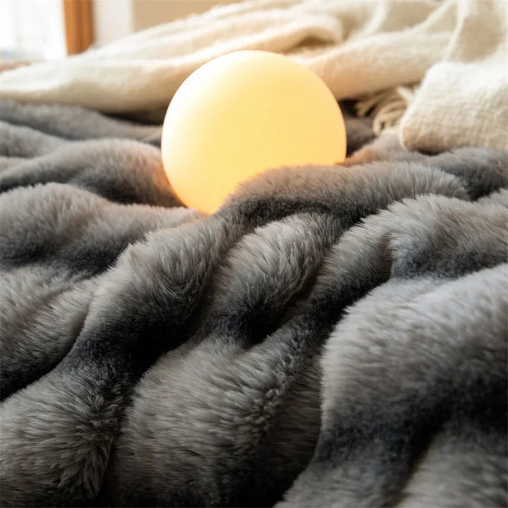A glowing round lamp sits on a plush, gray faux rabbit fur blanket with a luxurious throw featuring an elegant wave pattern in the background.