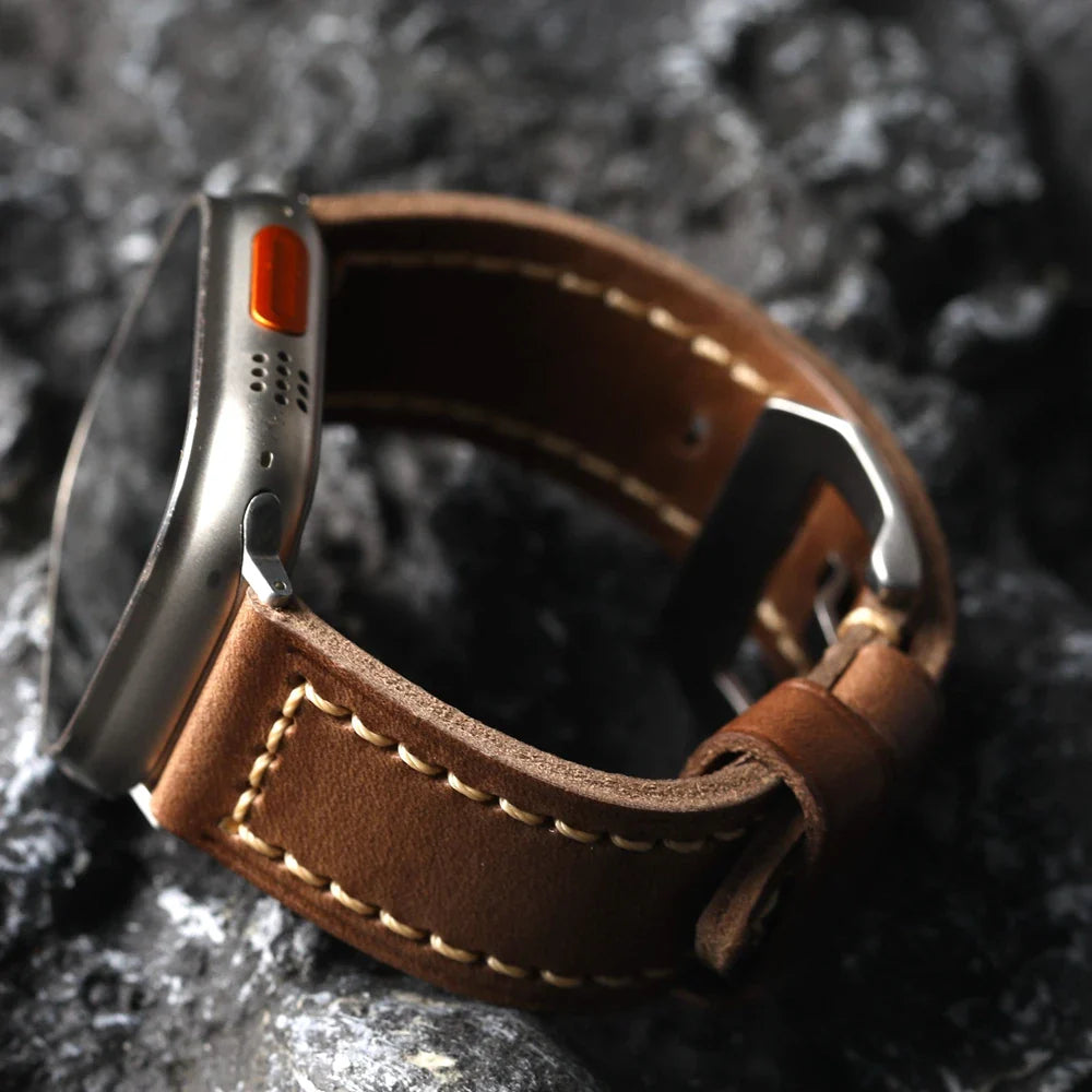 A close-up of a smartwatch with a handcrafted cowhide leather band and white stitching, placed on a dark, textured stone surface. The metallic case features an orange button on the side.