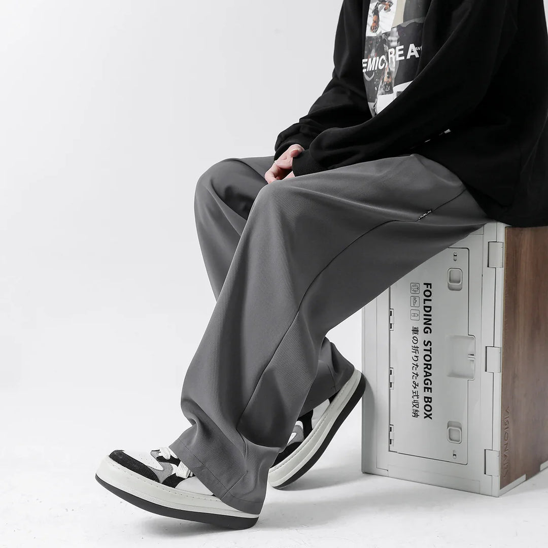 A person wearing loose gray streetwear joggers, black and white sneakers, and a black long-sleeve shirt sits on a light-colored storage box against a plain white background.