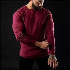 A man wearing a long sleeve fitted, moisture-wicking shirt and black pants stands against a dark background, looking down and adjusting his sleeve with one hand.
