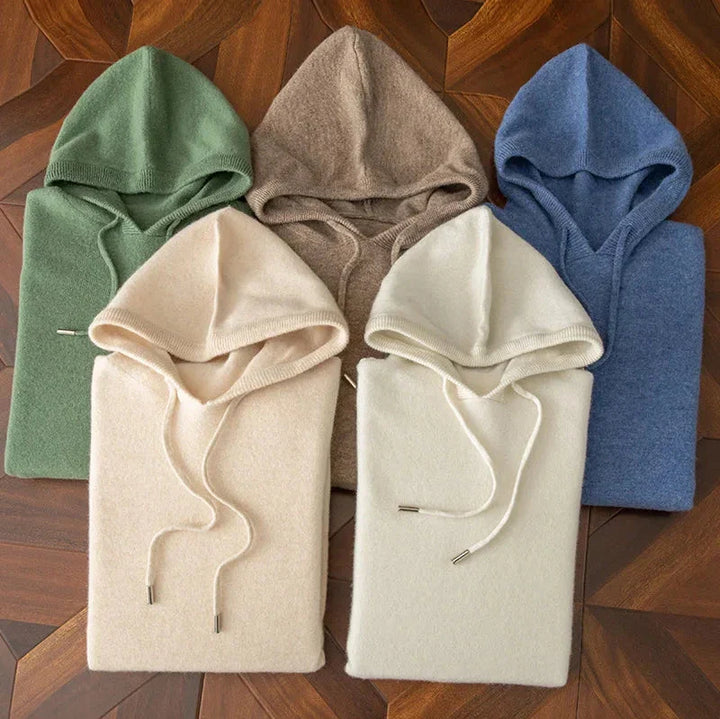 Five hooded sweaters in green, beige, blue, cream, and off-white are neatly folded and arranged on a wooden surface—the perfect layering sweater choices for a cozy look, with hoods and drawstrings stylishly visible.