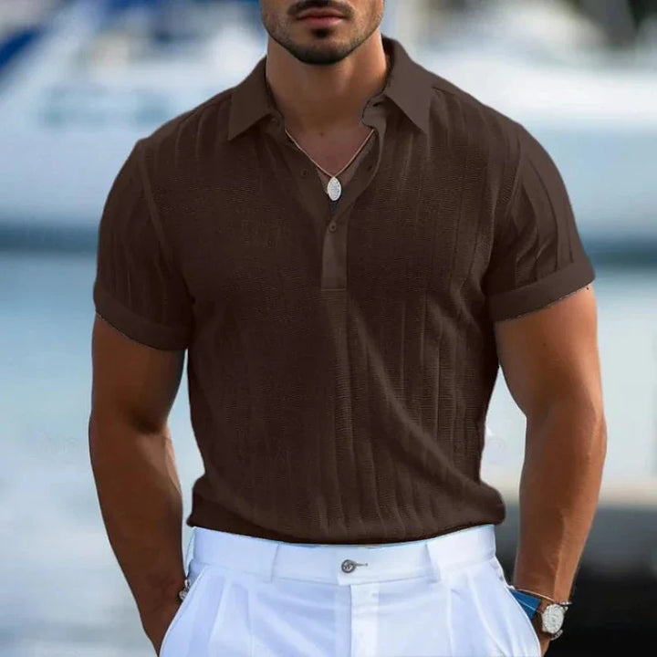 A man wearing a short-sleeved brown polo shirt in a tailored fit and white pants stands outdoors. He has a trimmed beard, a necklace, and rolled-up sleeves. The blurred background hints at an outdoor or waterfront setting.