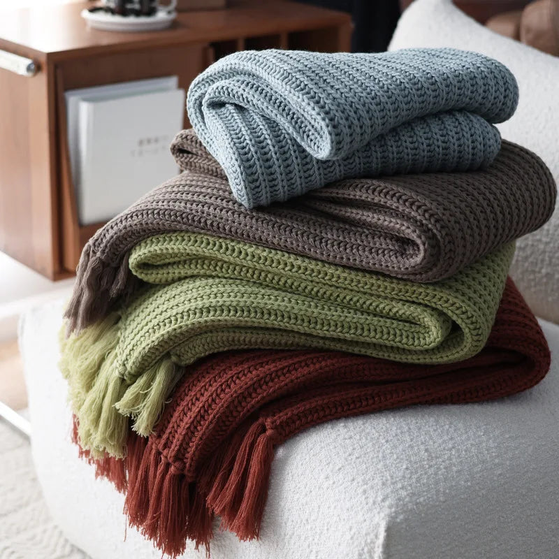 Four earth-tone throw blankets in blue, brown, green, and rust are neatly stacked on a white textured chair. A wooden cabinet with books and decorative items is visible in the background.