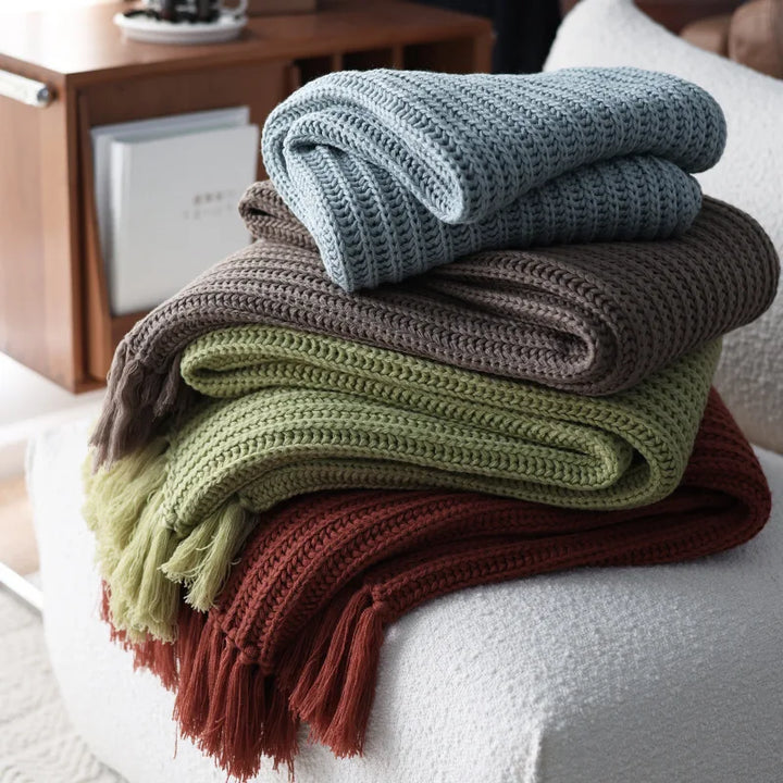 Four earth-tone throw blankets in blue, brown, green, and rust are neatly stacked on a white textured chair. A wooden cabinet with books and decorative items is visible in the background.
