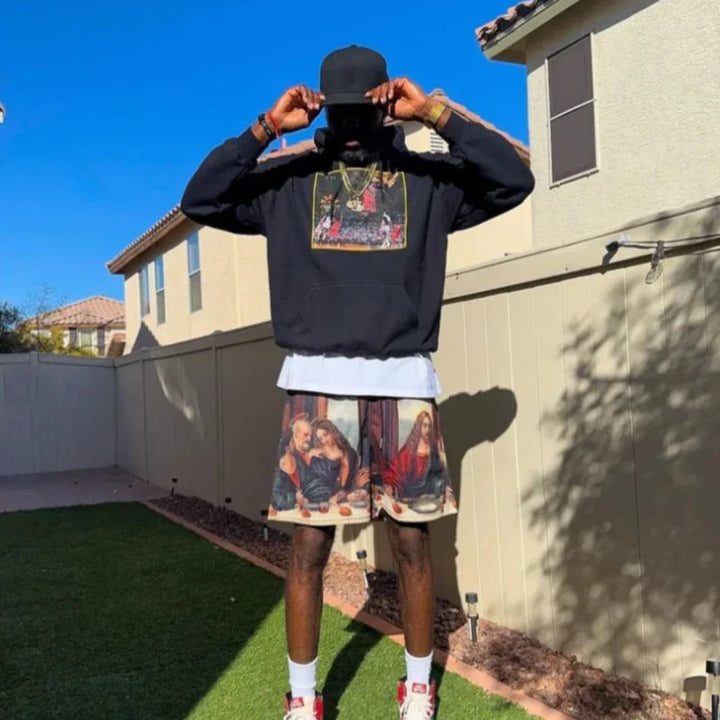 A person stands in a sunny backyard wearing a black cap, black hoodie with artwork, and Last Supper graphic shorts made from breathable mesh fabric. Their white t-shirt and sneakers with red accents complete the look, face hidden by the cap’s brim.