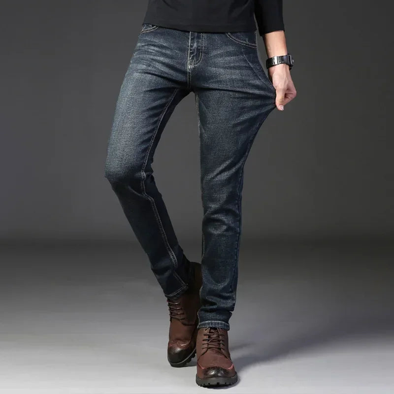 A person wearing dark blue stretch denim slim-fit jeans, brown lace-up boots, and a black top poses against a gray background, pulling the side of their jeans to show stretchiness.