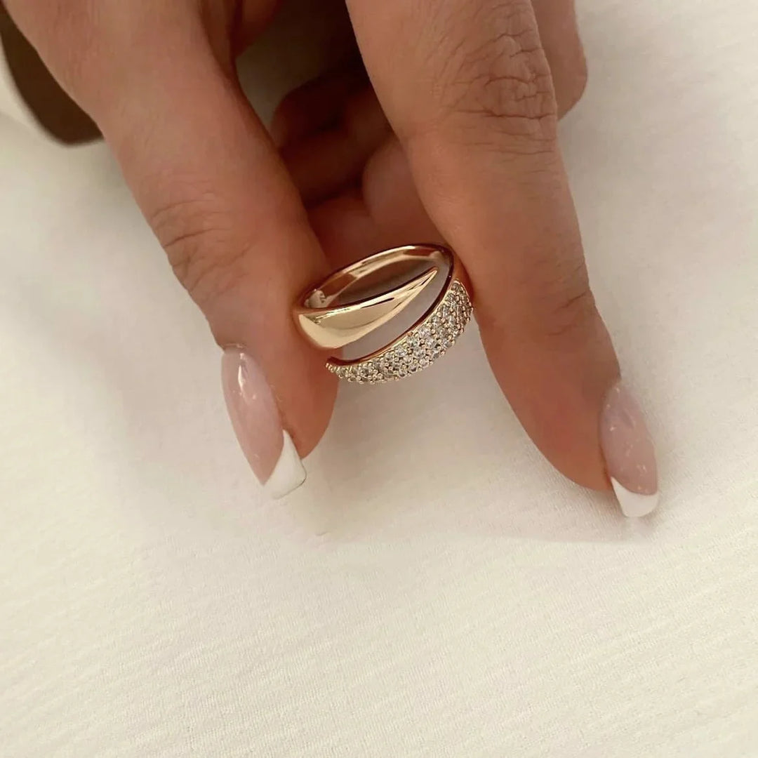 A hand with a French manicure holds a gold ring featuring a polished finish and pavé diamond accents, resting on a white fabric background.