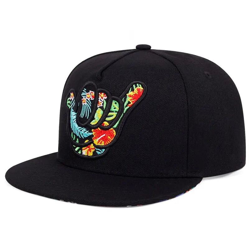 A black Hawaiian-inspired cap featuring an embroidered shaka hand sign adorned with vibrant tropical flowers and leaves in red, yellow, green, and blue on the front.