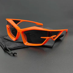 Futuristic cycling glasses with bright orange geometric frames and high-contrast, UV400 protection lenses are displayed on a black drawstring pouch against a dark background.