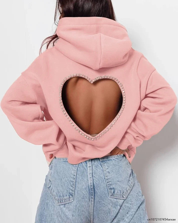 A person wearing a light pink oversized hoodie featuring a heart-shaped open back outlined with rhinestone trim, paired with high-waisted light blue jeans.