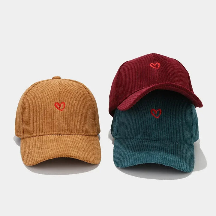 Three corduroy hats in mustard yellow, teal, and burgundy, each with a small red heart embroidery on the front and a comfortable FlexFit band, displayed against a white background.