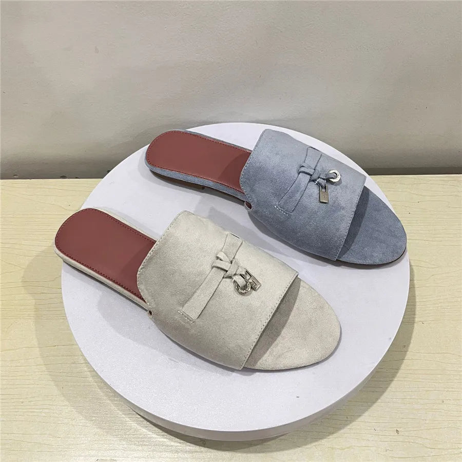 Two slip-on flat shoes with a suede finish are displayed on a round white platform. One is beige, the other light blue, and both feature a decorative bow with a small metallic charm on the front.
