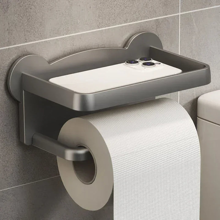 A modern wall-mounted toilet paper holder with a moisture-resistant, multi-functional shelf, holding a large roll and a white smartphone on top, is installed on a gray tiled wall next to a toilet.