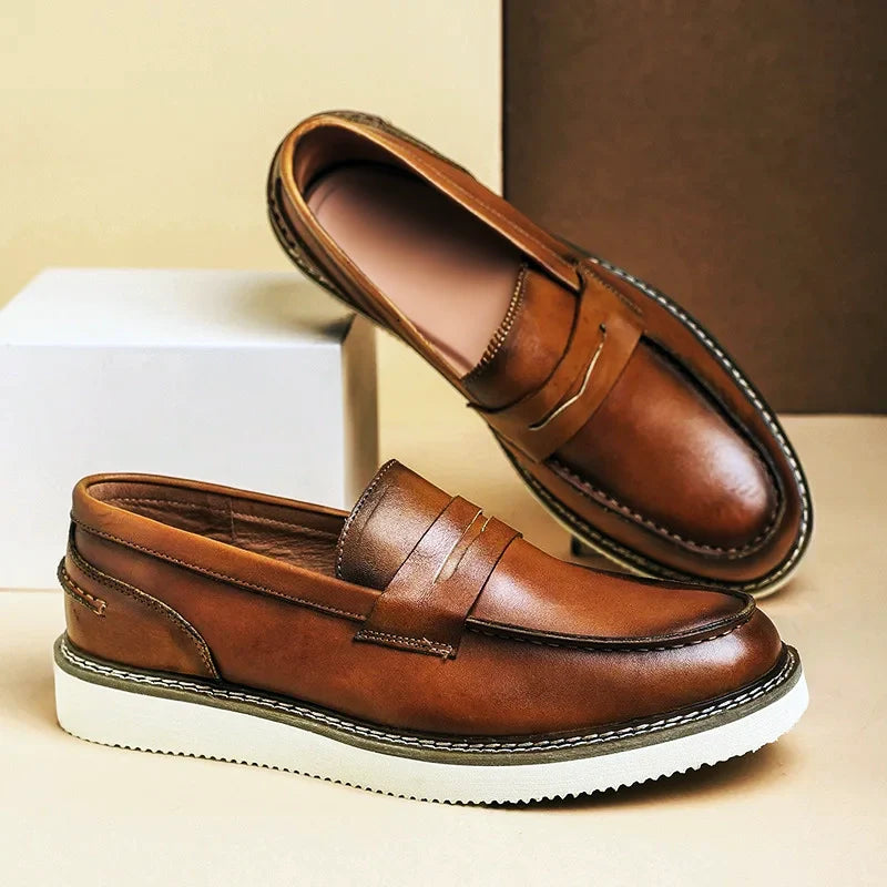 A pair of brown Italian leather penny loafers with white rubber soles, displayed against a beige and brown background. One shoe is upright, and the other is tilted, resting on a white rectangular block—perfect for business casual looks.