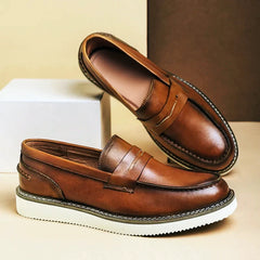 A pair of brown Italian leather penny loafers with white rubber soles, displayed against a beige and brown background. One shoe is upright, and the other is tilted, resting on a white rectangular block—perfect for business casual looks.