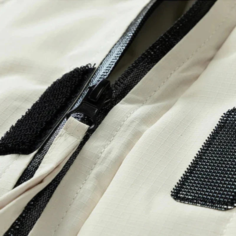 Close-up of a beige, windproof fabric with a black zipper, black Velcro strap, and a black mesh patch. The material has a subtle grid pattern and appears to be part of an insulated jacket or bag.