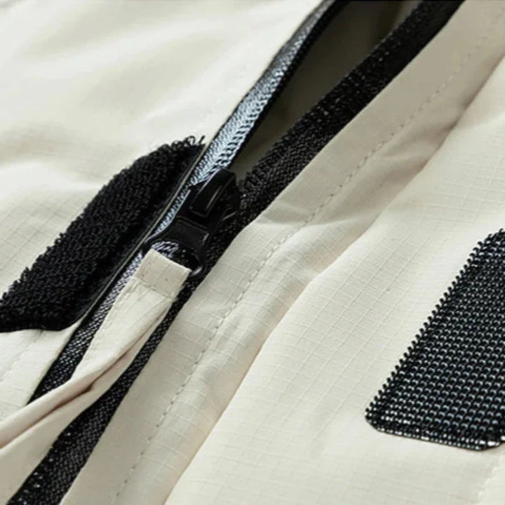 Close-up of a beige, windproof fabric with a black zipper, black Velcro strap, and a black mesh patch. The material has a subtle grid pattern and appears to be part of an insulated jacket or bag.