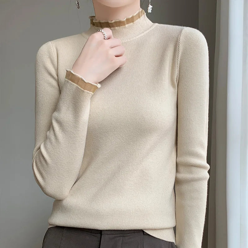 A person wears a fitted, long-sleeve beige sweater with a ruffled mock neck and wrist, made from a soft cashmere blend, paired with dark trousers. They stand near a light-colored curtain, one hand gently touching their neck.