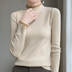 A person wears a fitted, long-sleeve beige sweater with a ruffled mock neck and wrist, made from a soft cashmere blend, paired with dark trousers. They stand near a light-colored curtain, one hand gently touching their neck.