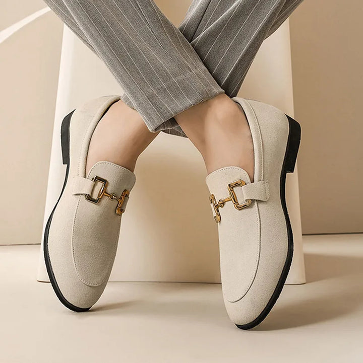 A person wearing luxury loafers with gold buckle details and black soles, paired with light gray pinstriped pants, sits with legs crossed against a neutral background.