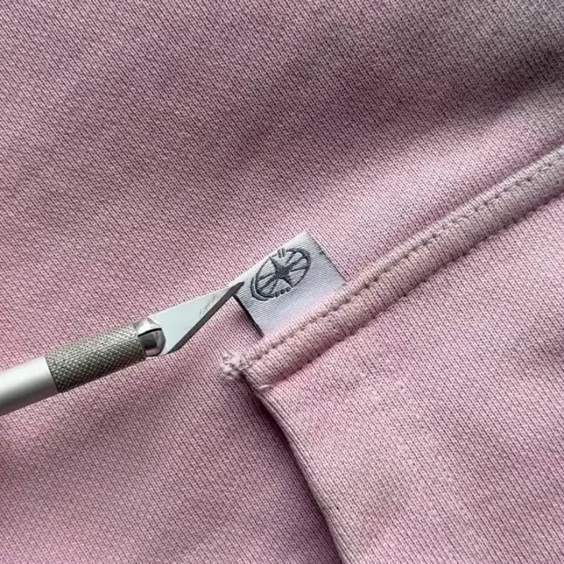 A craft knife points at a small white tag with a logo, sewn into the seam of a light pink garment—possibly a sweatshirt or hoodie—crafted from breathable fabric.