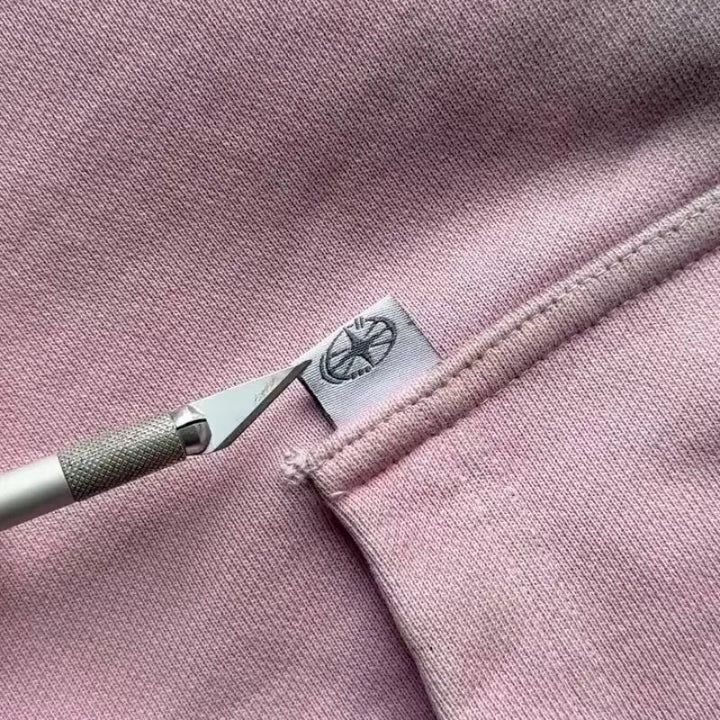 A craft knife points at a small white tag with a logo, sewn into the seam of a light pink garment—possibly a sweatshirt or hoodie—crafted from breathable fabric.
