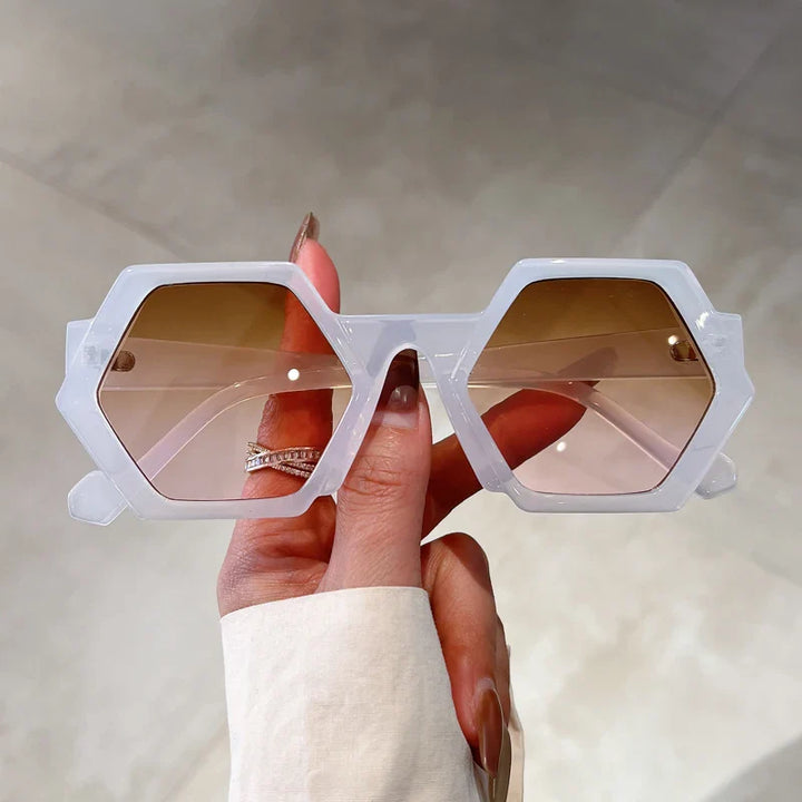 A hand with manicured nails and a ring holds up a pair of trend-forward, hexagonal frame sunglasses with white rims and brown gradient lenses, offering UV400 protection against a neutral background.