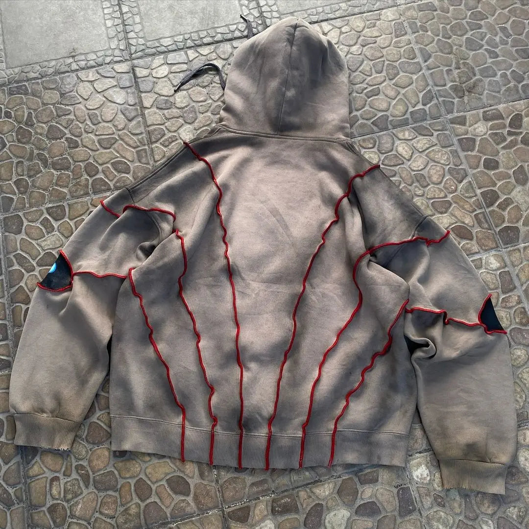 A brown hooded sweatshirt with red zigzag stitching and black patches on the elbows, laid flat on a stone-tiled surface. Crafted from breathable fabric, the stitching forms web-like patterns across the back and sleeves.